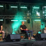 Na scenie od prawej: Tomasz Budzyński z gitarą, gitarzysta elektryczny, perkusista z kajonem.