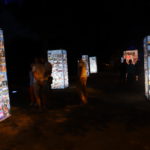 Wystawa zdjęć z okazji 50-cio lecia działalności MOK. Wieczór, ciemno, podświetlone konstrukcje ze zdjęciami, drzewa, widzowie.