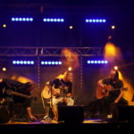 Na scenie siedzący Grzegorz Kupczyk z gitarą przed mikrofonem. Po lewej i po prawej dwóch siedzących gitarzystów z gitarami akustycznymi.