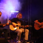 Na scenie siedzący Grzegorz Kupczyk z gitarą przed mikrofonem. Po lewej i po prawej dwóch siedzących gitarzystów z gitarami akustycznymi.